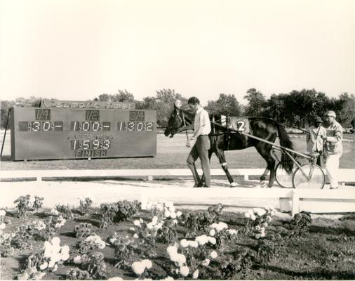 Horses; Harness Racing; Race Scenes; Men lead a horse past a flower garden and the timer