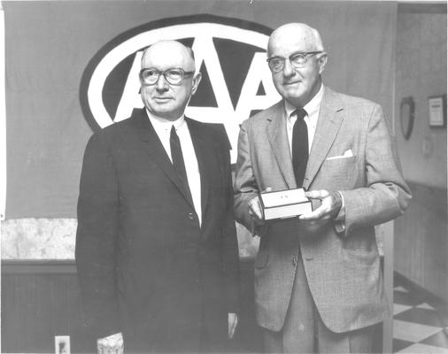 American Automobile Association; Two men, standing in front of a large AAA logo, with small gift box