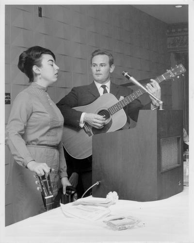 Musicians and Musical Instruments; A female singer and a male guitar player perform by a microphone