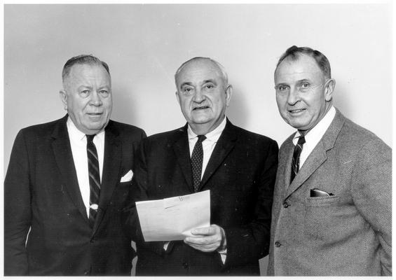 Rupp, Adolph F.; Rupp holds a stack of paper and stands between two men looking directly toward the camera