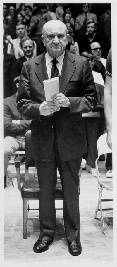 Rupp, Adolph F.; Rupp stands in front of a chair, staring toward the camera