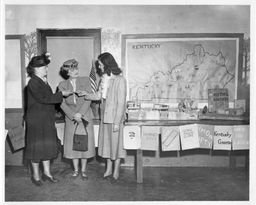 Women; Groups; Partially Identified; Three women stand by a table of children's projects