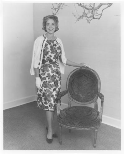 Women; Individual; Unidentified; A woman in a flowered dress stands by an antique chair