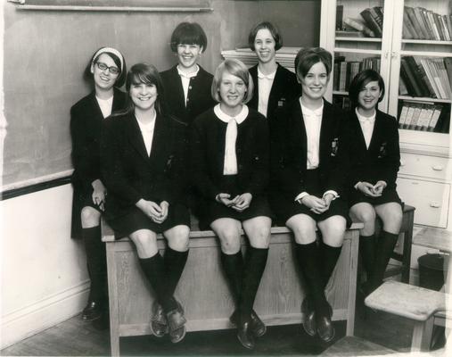 Groups; Unidentified; Seven teenage girls sitting on and standing near the teachers desk