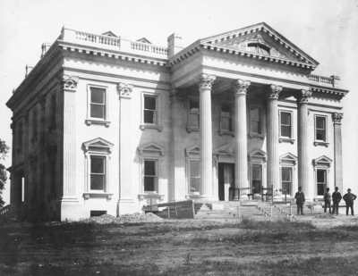 Exterior of Elmendorf Farm Mansion; a nearly completed version of the the construction from #10 and #11, four men are standing near the incomplete front steps, the building is a large, two-story mansion with tall columns on a front portico, there is still landscaping to be done. Silver Print