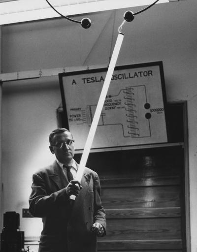 Yost, Francis L., Professor of Physics, pictured holding illuminated flourescent bulb, example of the Tesla oscillator shown in diagram in background