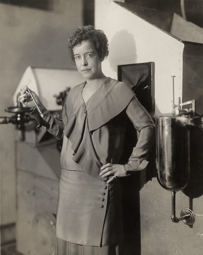 A portrait of Margaret Ingels holding a piece of machinery she invented. There is a tear on the bottom right side of the print