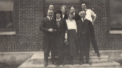 A group portrait of men and women on the steps of a building. The woman in white may be Eva Allen. This print was found pasted to the back of page 111 of 