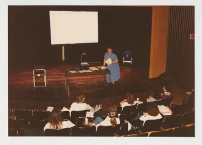 A female professor addresses a class meeting in a theater