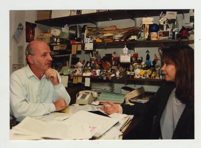 Professor Robert McAninch sits at the desk in his office while speaking with an unidentified woman