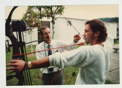 A male professor instructs a male student in archery