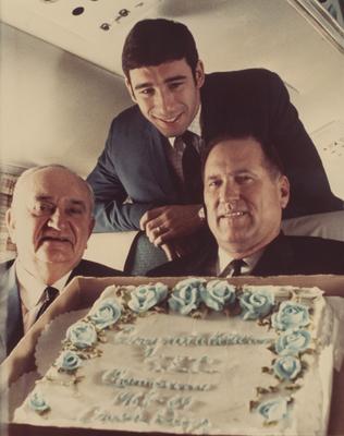 Coach Adolph Rupp (left), player Phil Argento (standing behind) and an unidentified man pictured on plane after being named the Southeastern Conference championships; they are holding a cake that reads 