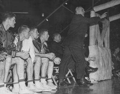 Basketball game action; photo of the bench, pictured left to right are Ed Beck, Vernon Hatton, two unidentified players, coach Adolph Rupp (standing) and assistant coach Harry Lancaster; photographer:  John Mitchell