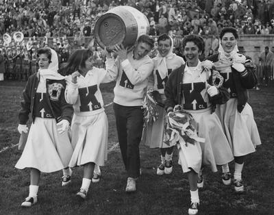 Five female cheerleaders with bobbie socks and saddle oxford shoes attending the homecoming football game at Stoll Field, November 23, 1957; pictured from left to right are Nancy Lowe, Pat Nallinger, Ellery 
