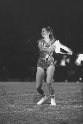 Unidentified majorette, using flaming batons, during band routine on the football field; game unidentified