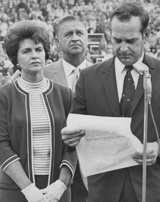 Dr. Claude Sullivan (right) reading from a paper while Athletics Director Harry Lancaster (center) and Alice Sullivan (left) stand and listen on the field during football game; Lexington Herald-Leader photo
