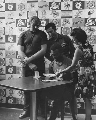 UK football players Ken Fuller (left), Darryl Bishop (center) and trainer Roy Don Wilson watch as Jean Ham, Director of Extension Service for Home Economics, demonstrates proper dining techniques; head coach Fran Curci (1973-81) invited the extension service to instruct the UK team in etiquette for dinners, banquets, etc.; Public Relations photo