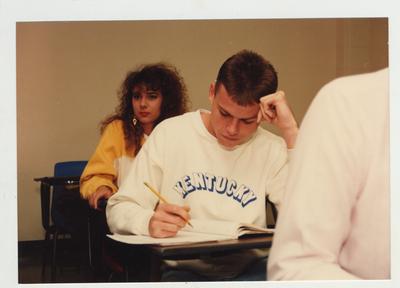 A male student takes notes and a female student listens during a lecture in a classroom