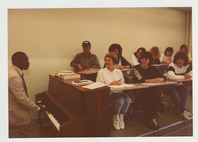 Students listen as a male African - American professor plays the piano in a classroom