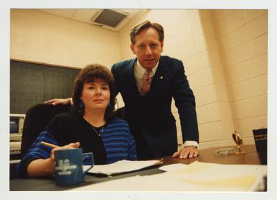 A man stands above a woman who sits at her desk