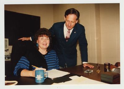 A man stands above a woman who sits at her desk
