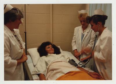 Female nursing students practice on a patient while a female professor (left) watches