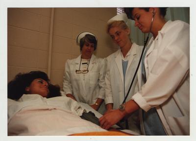 Female nursing students practice on a patient while a female professor (left) watches