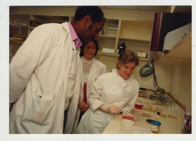 A woman and an African - American man watch a female student work in a laboratory