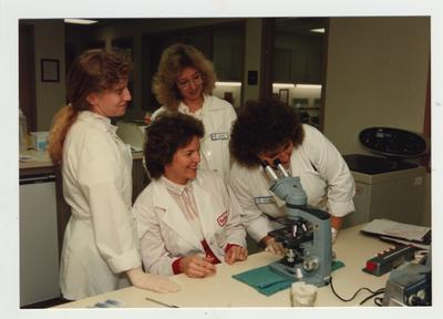 Woman watch as another woman looks through a microscope in a laboratory