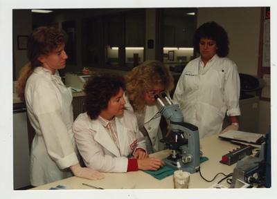 Woman watch as another woman looks through a microscope in a laboratory