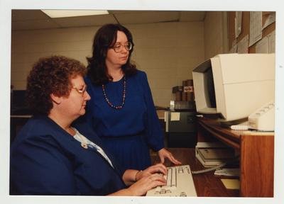 A woman stands near another woman typing on a computer