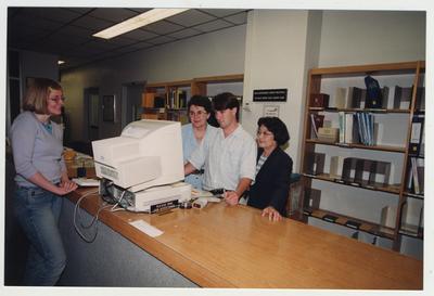 Librarian In Soon Chi (right), with an unidentified man and woman, standing behind the circulation desk looking at a computer and helping a female student in the Medical Library (located in the Medical Center); Photographer: Mark Minor