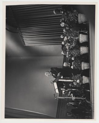 Dean William Willard (far right), Governor Bert Combs (second from right), President Frank Dickey (third from right), former President Herman Donovan (fourth from right), and others sit on a stage listening to an unidentified man speaking at the Medical Center dedication in Memorial Hall