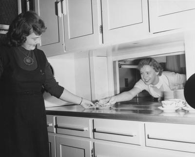 Two unidentified women serving and taking a plate of food in a practice house