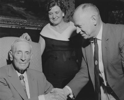 James W. Carnahan (seated, left) shaking hands with unidentified man as unidentified woman looks on, at reception for Carnahan House dedication