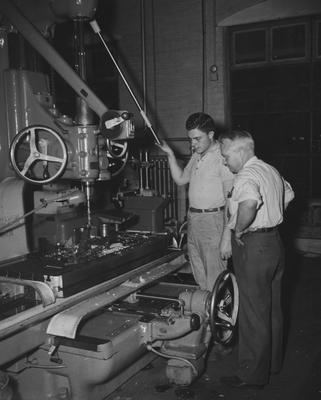 Two unidentified men are standing by a machine in the engineering department