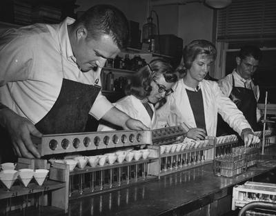 Four students performing an experiment. From left to right: unidentified man, unidentified woman, Gloria Sawtelle, unidentified man. Received on March 31, 1961 from Public Relations