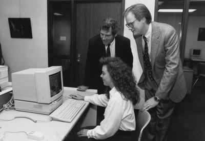Mark Denomme (standing to the right) and two unidentified people gather around a computer in the computer lab for this photograph