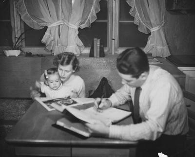 The Lawyer-Brock  family is seated at a table in a prefabricated Cooperstown house. From left to right: Kathie Brock, Mrs. Brock, and Mr. James C. Brock (a law student)