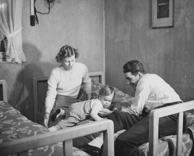 The Lawyer-Brock  family is in the bedroom of a prefabricated Cooperstown house. From left to right: Mrs. Brock, daughter Kathie Brock, and Mr. James C. Brock (a law student). Mr. Brock was a University of Kentucky student veteran, able to live with him family in the Cooperstown Veteran's Housing Project