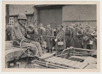 Elmer Ayers of Lexington Kentucky, Ninth US Army, sits beside his machine gun mounted on a jeep while German prisoners are lined up.  Stendal, Germany