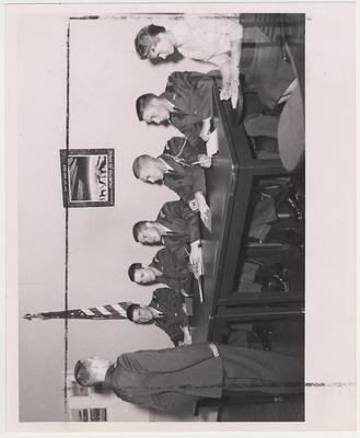 An officer or cadet standing at attention in front of the board.  In the center is John Zackerman