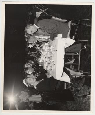 President Frank Dickey (left, facing the camera) is sitting at a table with his family.  Mildred (spouse of Governor Chandler) is the first person on the left, Governor A. B. 