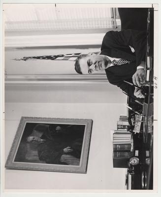 President Frank Dickey seated behind his desk with a portrait print of President Donovan on the wall to the left