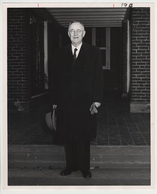 President Donovan standing in a top coat with his hat and gloves in hand