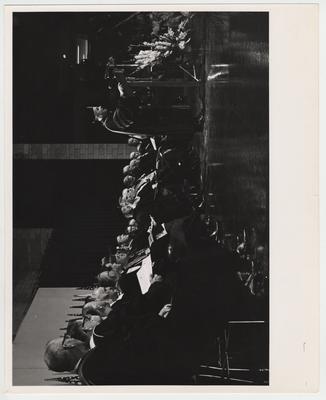 President John Oswald is standing at a podium, speaking at his inaugural ceremony.  Former president Frank Dickey (seventh from the right) is sitting on stage