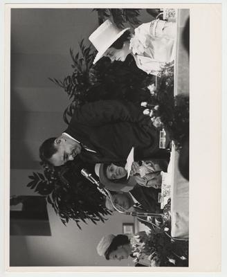 President John Oswald is standing and speaking at his inaugural luncheon in the ballroom of the Student Center.  His wife, Rosanel Oswald, is seated to president Oswald's left, and Governor Ned 