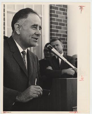 President Oswald (left) is speaking behind a podium.  In the background is William (Bill) Gant (right with arms folded), a member of the University of Kentucky Alumni Association Executive Committee