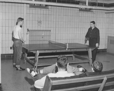 Two unidentified men are playing ping pong and two unidentified men are watching, in the basement of a boy's dorm