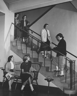 Two unidentified women are seated by stairs and four unidentified women are going up and down stairs in Jewell Hall. Jewell Hall was named after Mary Frances Jewell. Received March 16, 1957 from Public Relations